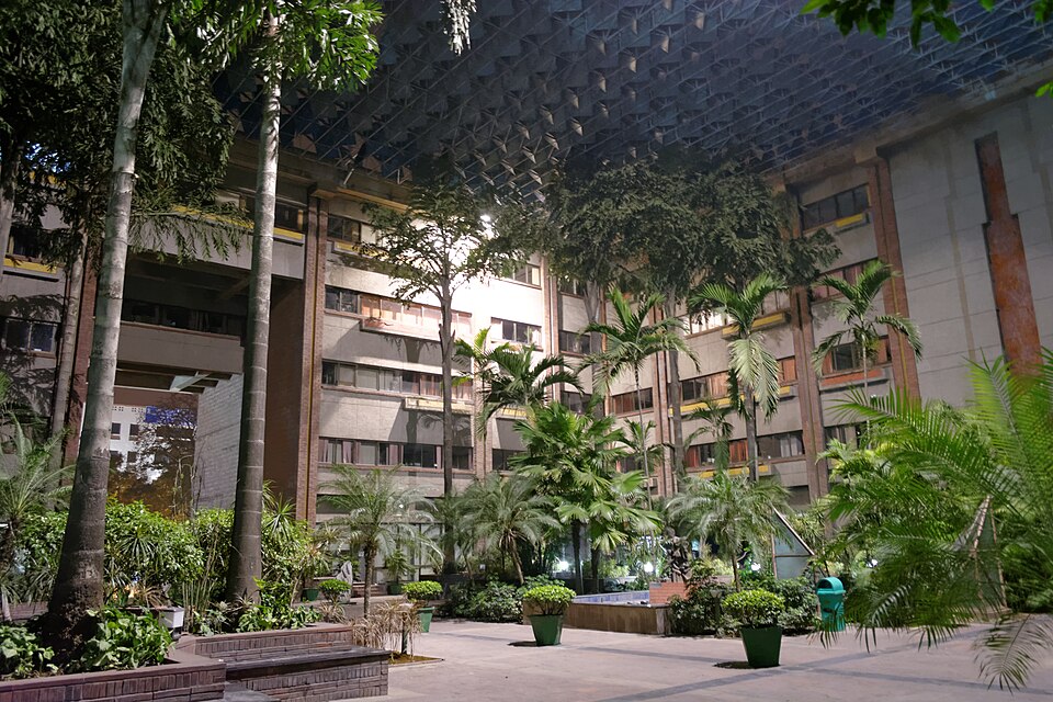A planted internal courtyard of the India Habitat Centre on Lodhi Road, New Delhi, with palms and shrubs under the complex's blue steel space-frame sun canopy
