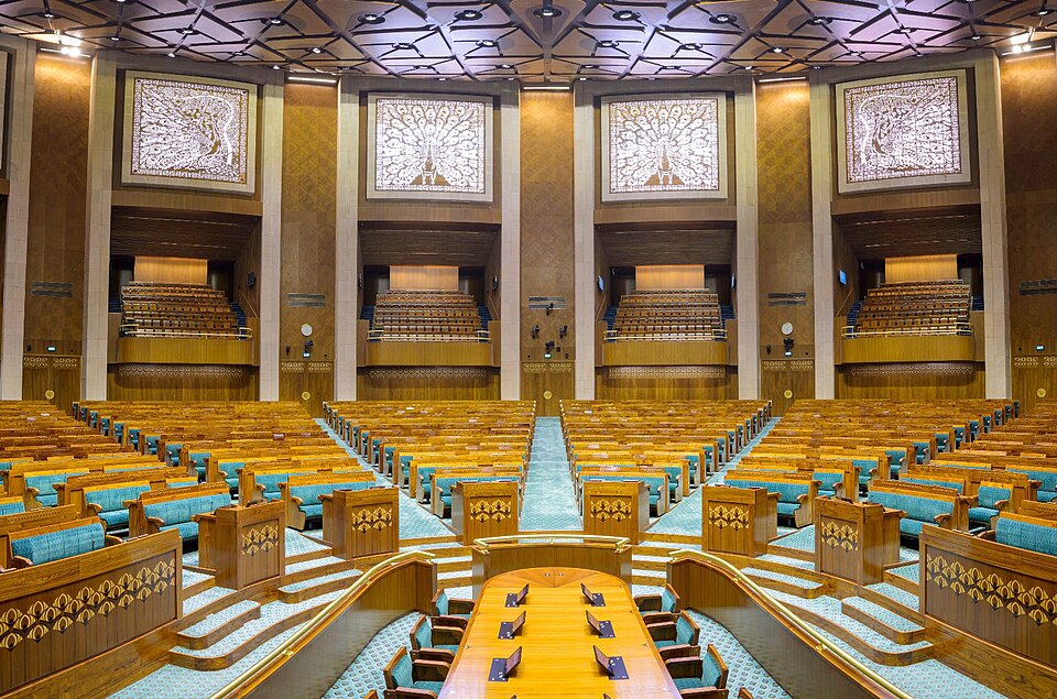 Interior of the Lok Sabha chamber in the new Parliament building, New Delhi, showing the fan of tiered wooden members' benches with teal upholstery, the Table of the House in front and peacock-motif screens above the galleries