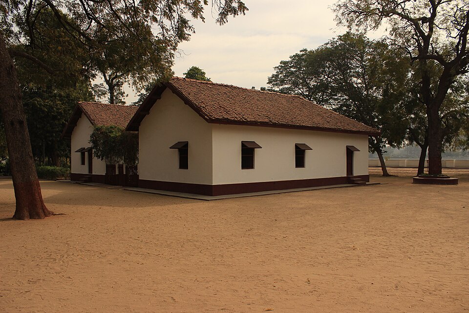A whitewashed, tile-roofed ashram building with shuttered windows at Sabarmati Ashram, Ahmedabad, standing on bare sandy ground among trees near the Sabarmati river