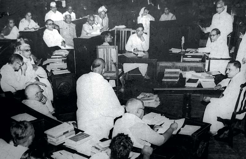 Black-and-white photograph of the Constituent Assembly of India in session, members in white khadi seated at desks piled with papers and bound volumes while one member speaks from a raised desk