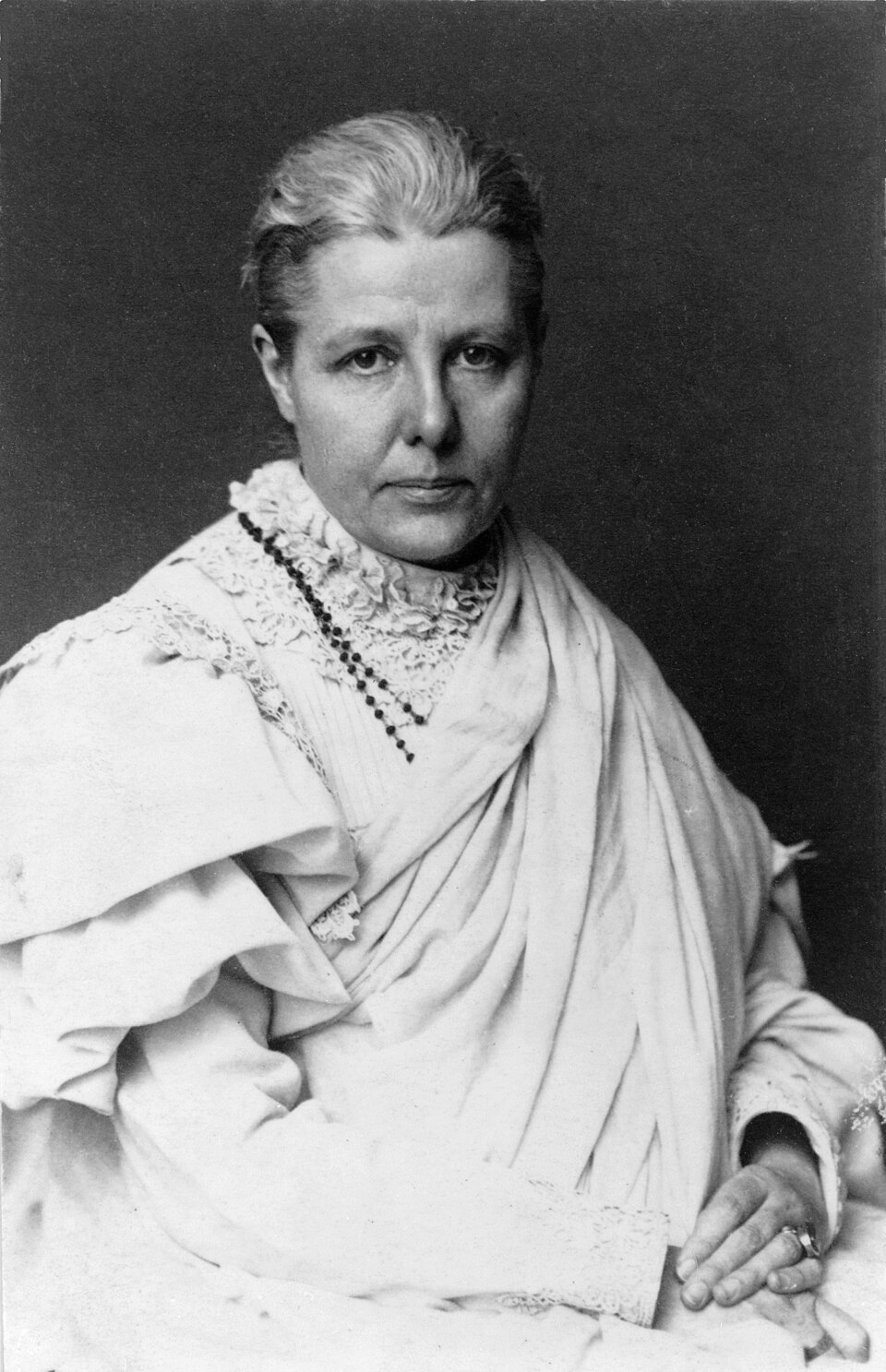 Studio portrait of Annie Besant in later life, seated in a white shawl and lace-collared dress with a long beaded necklace