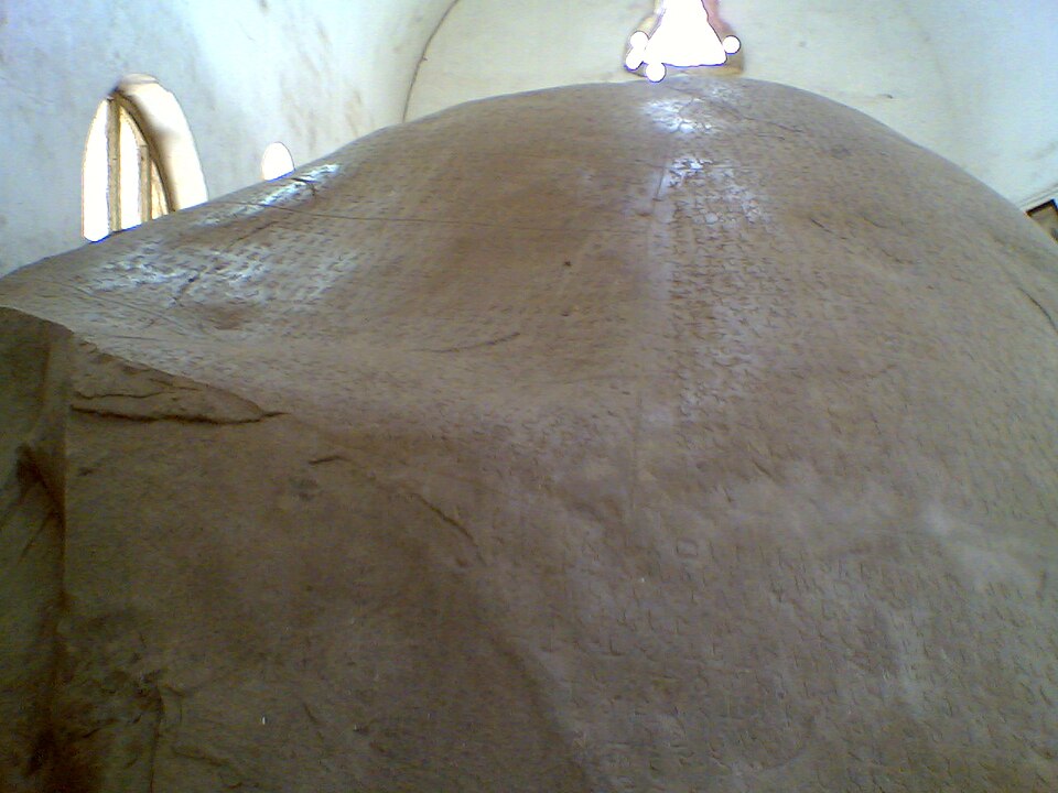 The large domed granite boulder carrying Ashoka's Major Rock Edicts at Junagadh, its surface covered in rows of shallow Brahmi characters, housed inside a whitewashed protective shelter