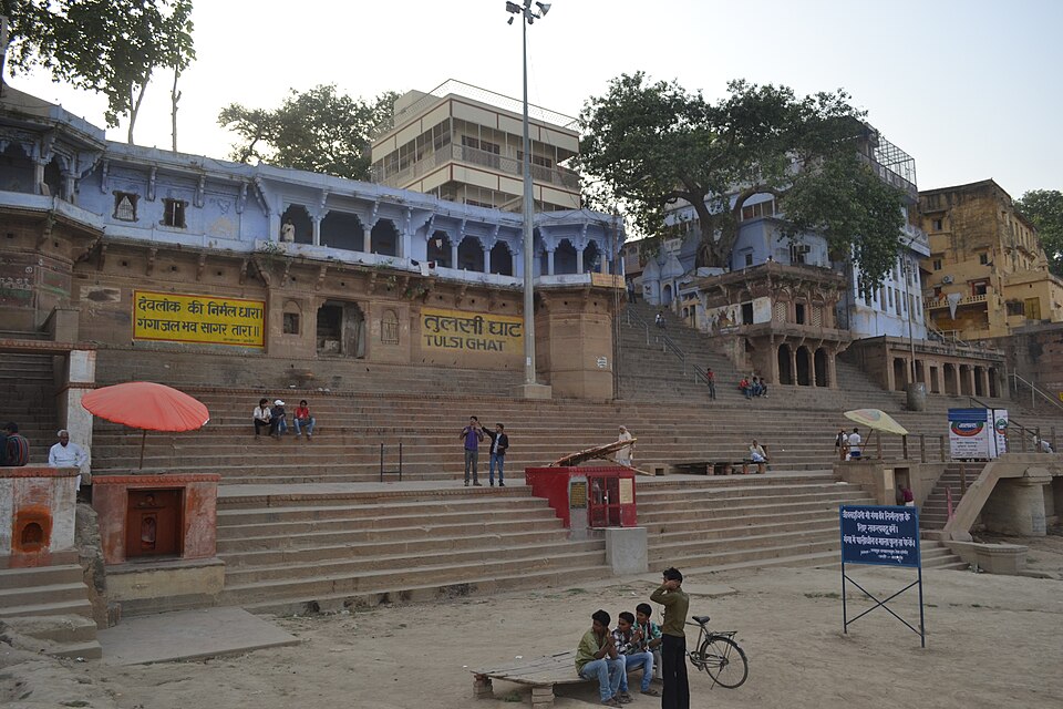 The wide stone steps of Tulsi Ghat at Varanasi rising from the Ganga to a pale blue arcaded building, with the name TULSI GHAT painted in Devanagari and Latin letters on the ghat wall