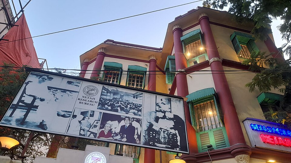 Netaji Bhawan, the Bose family house on Elgin Road in Kolkata, a cream-and-red three-storey building with green louvred shutters, a Netaji Research Bureau display board and neon sign at the entrance
