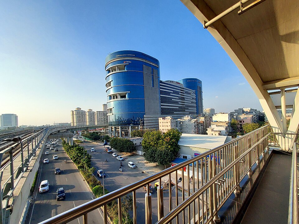 A curved blue glass office tower carrying American Express and goibibo signs stands above a multi-lane highway, with an elevated metro viaduct running alongside and low houses spreading behind.