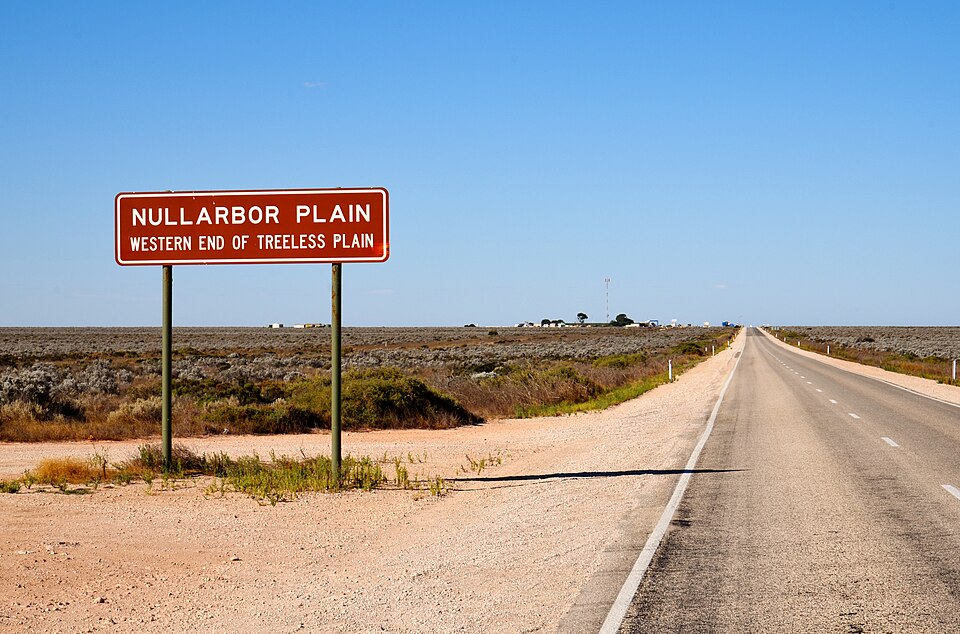 A dark red roadside sign reading 'Nullarbor Plain, western end of treeless plain' stands on the sandy verge of a straight two-lane highway that runs to a flat horizon across an endless expanse of low grey-green scrub under a clear blue sky.