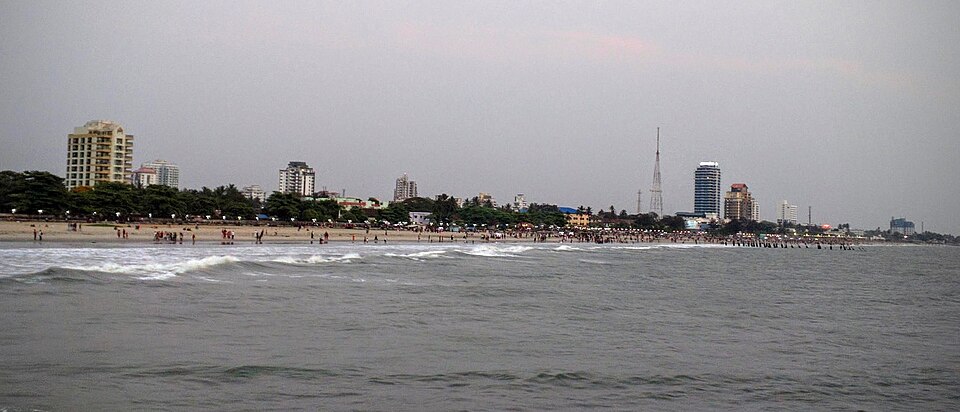 A city seafront seen from out at sea: choppy grey-green water fills the foreground, breaking in low white lines onto a pale sand beach where small groups of people stand at the water's edge, a row of dark wooden pier stumps runs out into the surf on the right, and behind the beach a fringe of coconut palms gives way to apartment blocks, a blue-glass tower and a tall lattice mast under a flat grey sky.