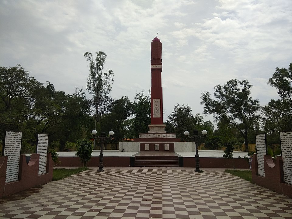 A tall red memorial pillar on a stepped plinth at the end of a checkerboard-paved walkway, with inscribed stone panels on low walls to either side.