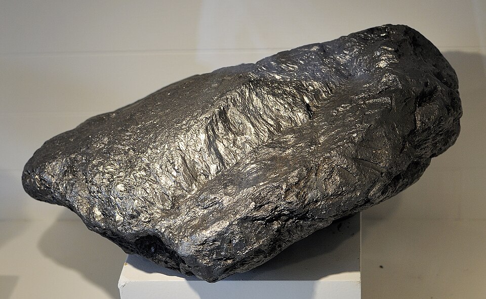 A large silvery-grey mineral block with a metallic sheen and a coarse, layered, splintery surface, resting on a white plinth against a plain pale background.