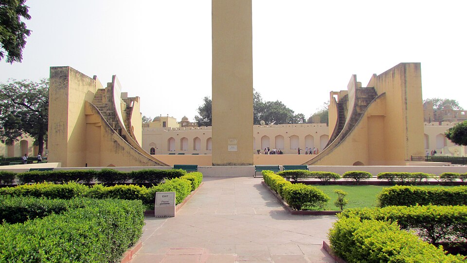 A tall yellow masonry wall rises straight up in the middle of a paved courtyard, flanked by two symmetrical curved stone ramps marked with steps; clipped hedges line the walkway in front and visitors stand under an arcade behind.