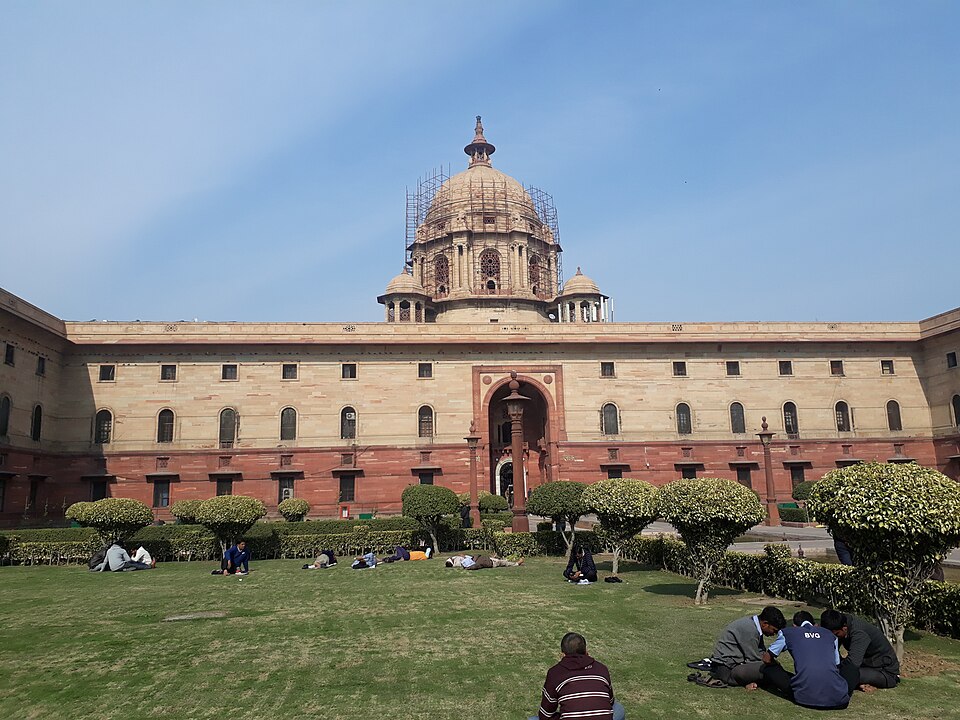 A long two-storey government building of cream and red sandstone with rows of arched windows and a central arched doorway, topped by a stone dome wrapped in scaffolding; students sit on the lawn among clipped round trees in front.