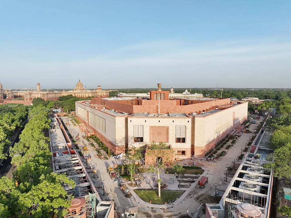Aerial view of a large triangular sandstone building with pale stone walls, tall shuttered windows and a bronze four-lion emblem mounted on its roof, ringed by construction hoardings, cranes and bare earth, with dense green treetops on both sides and a domed building flanked by long colonnaded blocks on the distant skyline.