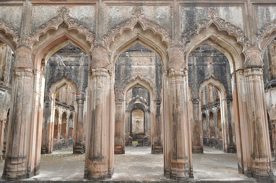 Rows of tall pointed cusped arches on thick plastered columns inside a roofed but derelict hall; the carved stucco above the arches is broken and stained black, and an empty arched niche closes the far end.
