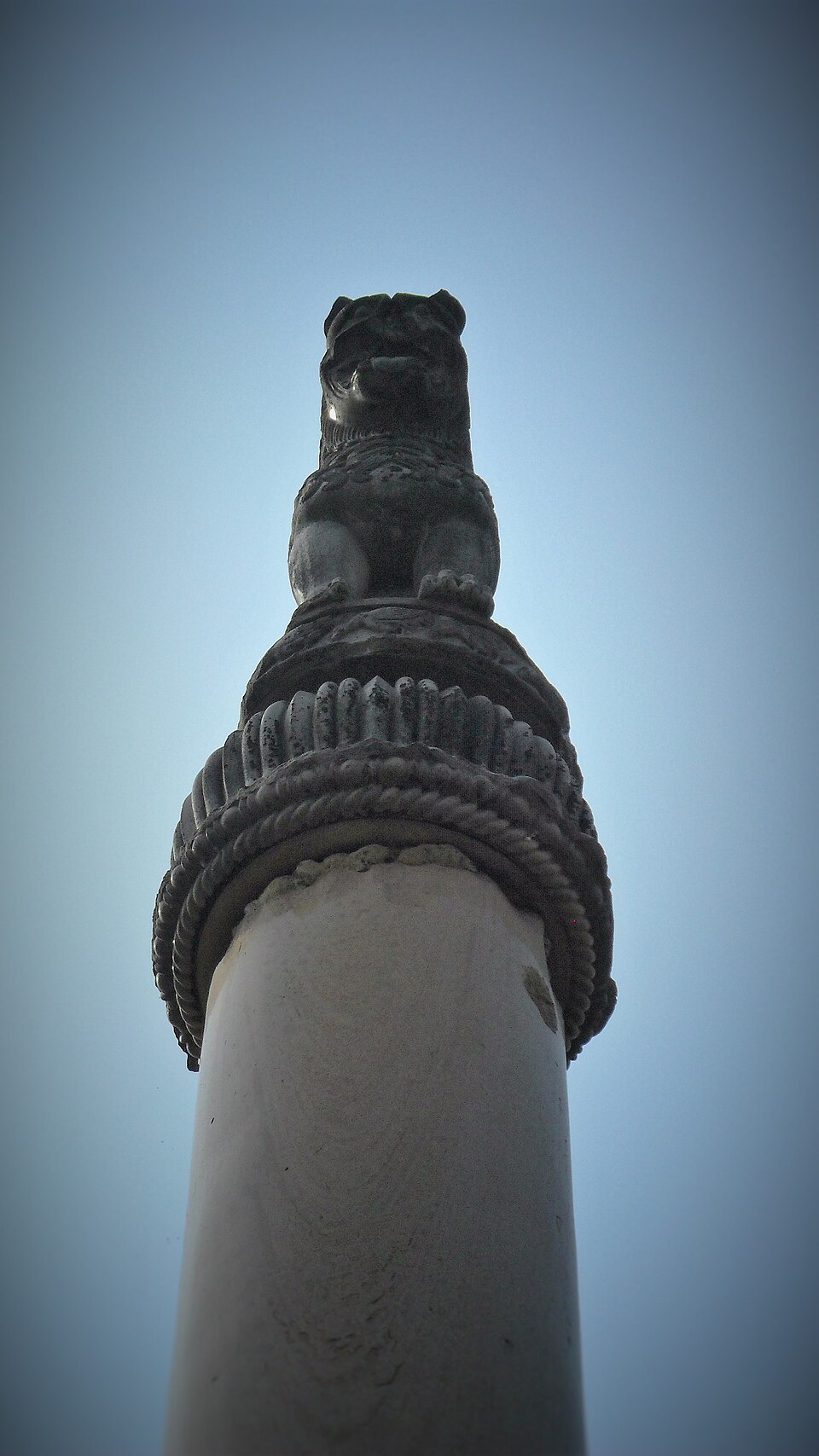 Seen from ground level looking straight up, a stone lion sits on a bell-shaped carved capital with beaded bands, crowning a smooth pale monolithic column against a plain sky.