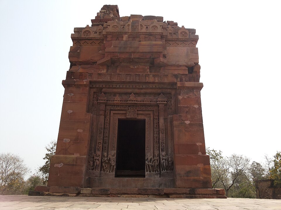 पत्थर की पक्की जगती पर खड़ा लाल बलुआ पत्थर का वर्गाकार देवालय, जिसका अँधेरा खाली द्वार खड़ी आकृतियों से गहरे तक्षित द्वार-स्तंभों और तक्षित द्वार-पट्टी से घिरा है, पीछे टूटा हुआ सीढ़ीदार शिखर उठा है और दोनों ओर सूखी झाड़ीदार वनस्पति है।