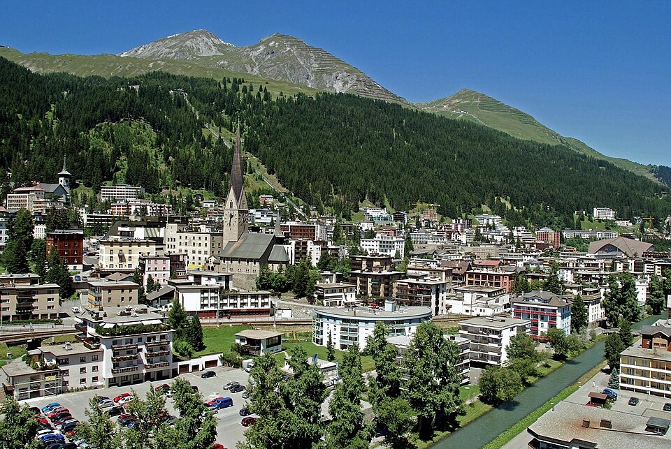 A Swiss valley town photographed from above in summer: dense blocks of hotels and chalets along the valley floor around a stone church with a tall pointed spire, a steep forested mountainside behind cut by pale ski runs and a lift line, and bare rocky peaks against a clear blue sky.