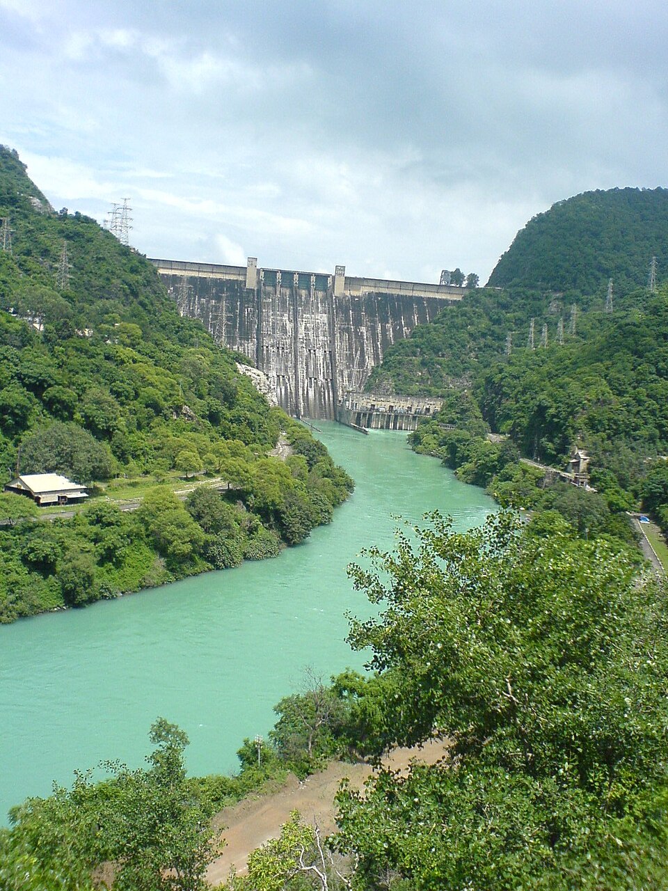 A tall grey concrete dam wall spans a narrow gorge between steep forested green hills, a turquoise river curving away from powerhouse buildings at its base, with electricity pylons along the ridge lines under a cloudy sky.
