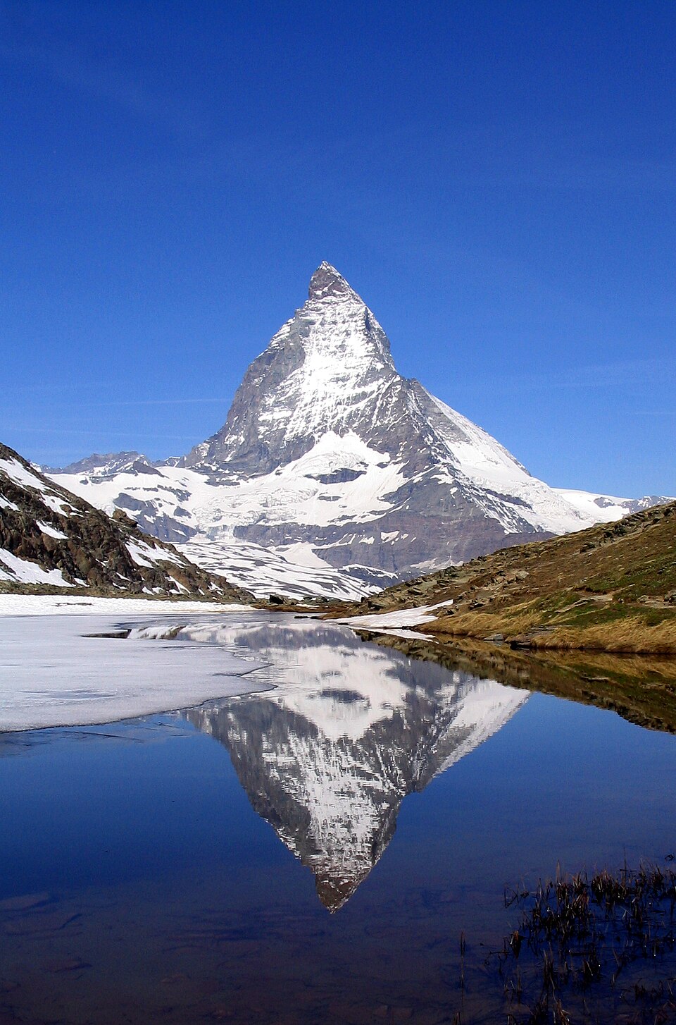 A steep snow-covered rock pyramid with a hooked summit rises against a deep blue sky and is mirrored in a partly frozen mountain lake in the foreground.