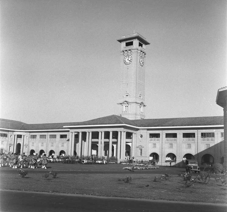 A black-and-white photograph of a long single-storey colonnaded government building with a tall square clock tower rising above its centre, a uniformed band lined up on the lawn in front and a vintage car parked to the right.