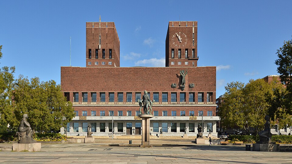 A wide brick civic building with two tall square towers, a clock set high in the right-hand tower, bronze figures mounted on the facade, and a paved open square with statues and trees in the foreground.
