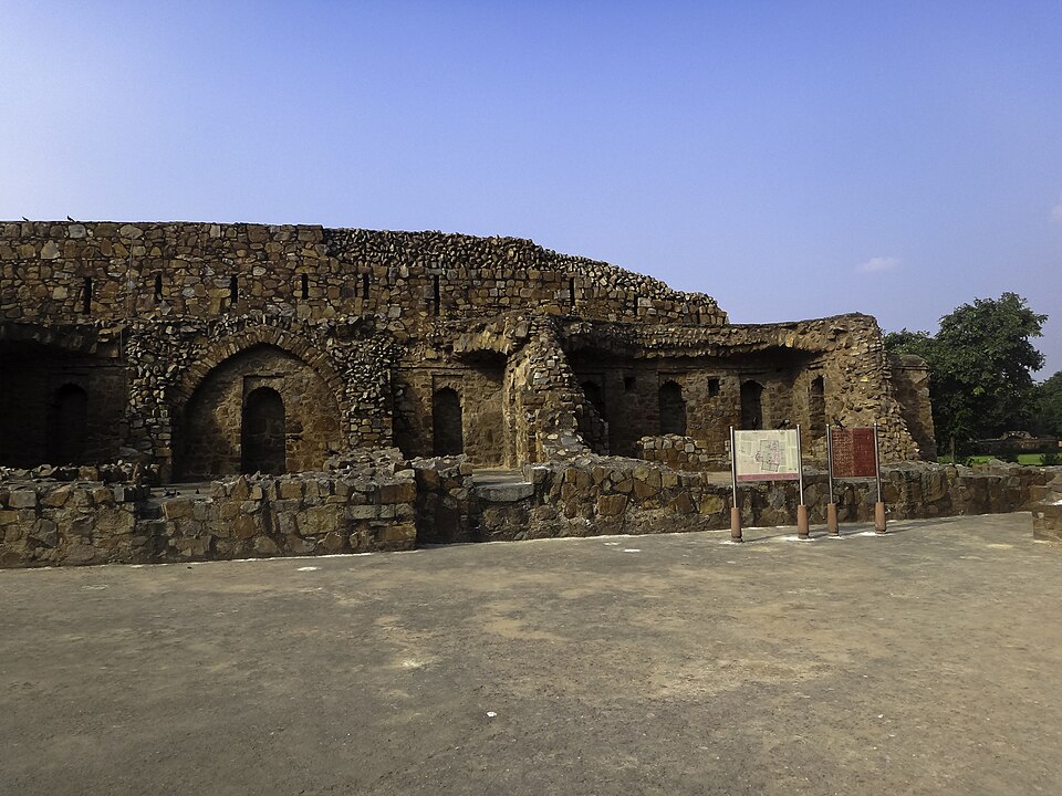 खुरदुरे पत्थर की चिनाई वाली एक लम्बी दीवार, जिसमें नुकीले मेहराबदार आलों और छोटे द्वारों की एक पंक्ति है, जिसका एक भाग ढहकर ढीले पत्थरों के ढेर में बदल गया है और जो एक नीची कुर्सी के पीछे खड़ी है; खुले नीले आकाश के नीचे बजरी भरे सूने प्रांगण में खम्भों पर लगे दो सूचना-पट्ट खड़े हैं, जिनमें से एक पर स्थल-मानचित्र छपा है।