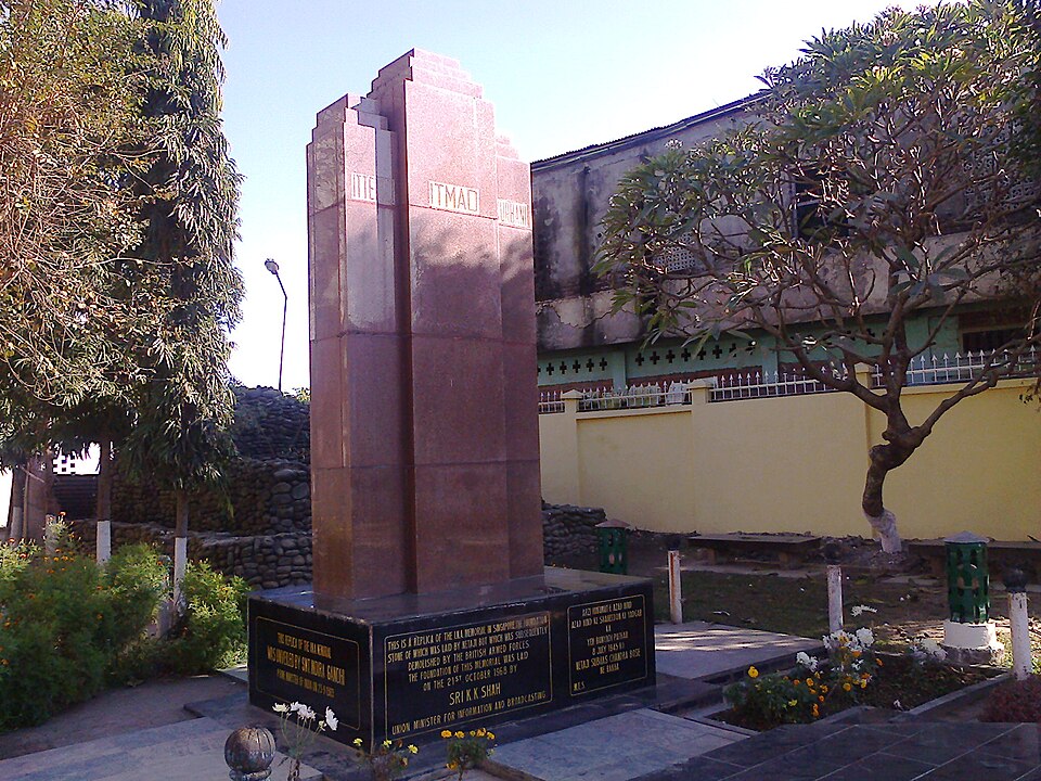 A stepped reddish-stone memorial pillar carrying carved words on its face, standing on a black granite plinth with inscribed plaques, in a small walled garden with flowering beds and a pale green building behind.