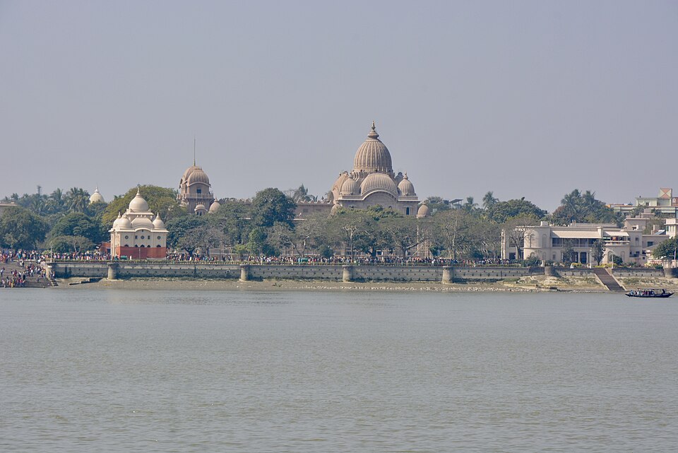 A broad grey river fills the foreground; on the far bank a pale sandstone temple with a large ribbed dome stands among trees, with a smaller white domed shrine to its left and other spires behind, all above a long stone embankment crowded with people, and a small boat at the right-hand bank.