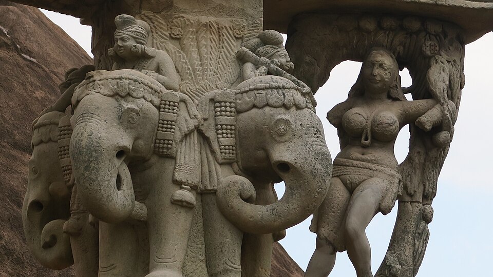 अपक्षयित बलुआ पत्थर की नक़्क़ाशी का निकट दृश्य : स्तंभ-शीर्ष के रूप में कंधे से कंधा मिलाकर खड़े तीन अलंकृत हाथी, जिनकी गर्दन पर सवार बैठे हैं, और दाहिनी ओर एक स्त्री-आकृति, जो एक हाथ उठाए घुमावदार पत्थर के कोष्ठ के नीचे से बाहर की ओर झुकी है।