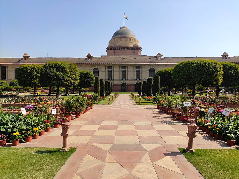 बलुआ पत्थर का लंबा प्रासाद, जिसके मध्य में धारीदार गुंबद और उस पर भारतीय तिरंगा है, और जो एक शतरंजी पत्थर-पथ के आर-पार दिखता है — पथ के दोनों ओर छँटे हुए शंक्वाकार वृक्ष तथा औपचारिक उद्यान में गमलों में लगे फूलों की पंक्तियाँ हैं।
