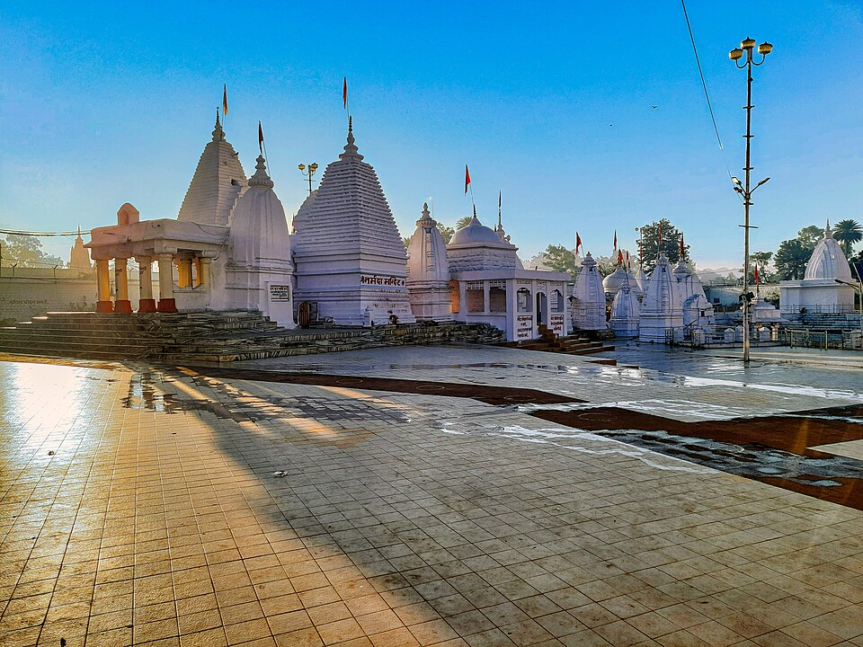 सफ़ेदी पुते पत्थर के मंदिरों का समूह, जिनके शिखर मंज़िलदार हैं और ऊँचे खंभों पर केसरिया ध्वजाएँ लगी हैं; ये सब सुबह की तिरछी धूप में साफ़ नीले आकाश के नीचे एक ख़ाली, गीले टाइल वाले प्रांगण के चारों ओर खड़े हैं।