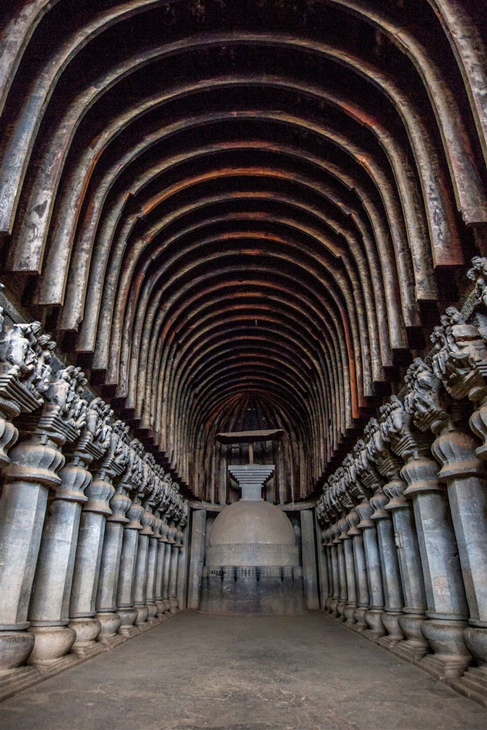The interior of the Great Chaitya at Karla, a rock-cut hall with a ribbed barrel-vaulted ceiling, twin colonnades of carved pillars and a stupa at the far end