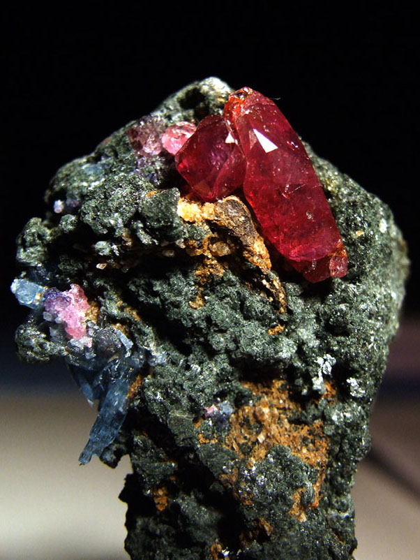 Deep red ruby crystals embedded in dark green rock matrix, with smaller pink and blue corundum crystals alongside