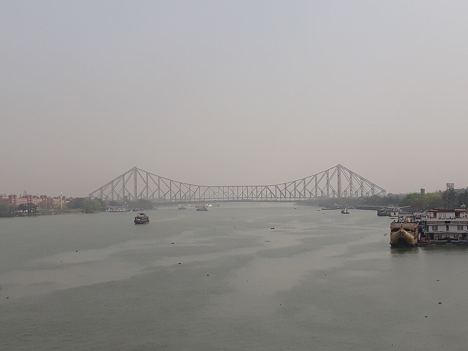 The full cantilever span of the Howrah Bridge across the Hooghly at Kolkata, with launches on the water and a moored river vessel at the near bank