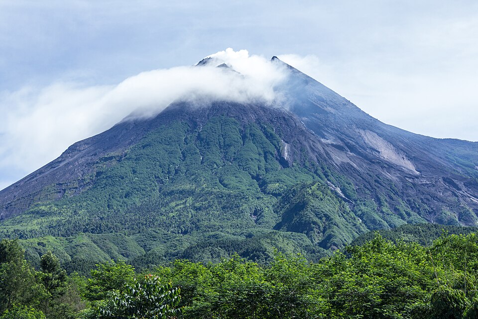 Mount Merapi's forested stratovolcano cone rising to a steaming summit wreathed in cloud, grey ash and lava scars streaking its upper flanks