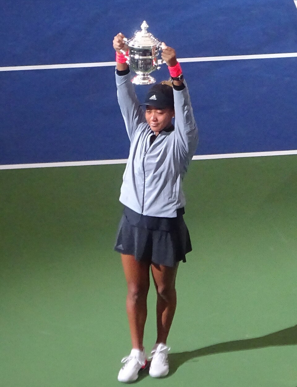 Naomi Osaka standing on a hard court, holding the US Open singles trophy raised above her head