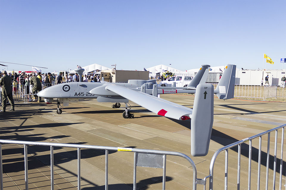 An IAI Heron unmanned aircraft on static display: a slender high-aspect-ratio wing, bulbous nose radome, twin tail booms and a rear pusher propeller, standing behind crowd barriers at an airshow