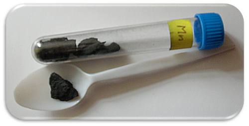 A specimen vial labelled Mn holding dark grey lumps of manganese metal, with one lump laid out on a white spoon beside it