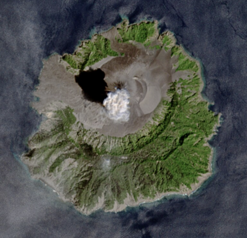 Satellite view of Barren Island in the Andaman Sea: a central ash-grey cone with a summit crater venting a white plume, ringed by dark lava flows and green vegetation, the island surrounded by open sea