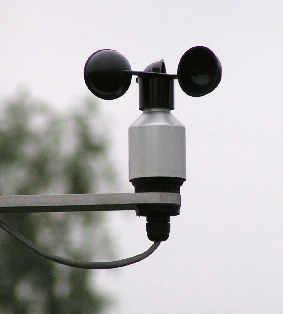 Close-up of a three-cup anemometer on a bracket, its black hemispherical cups mounted on a rotating spindle above the sensor body