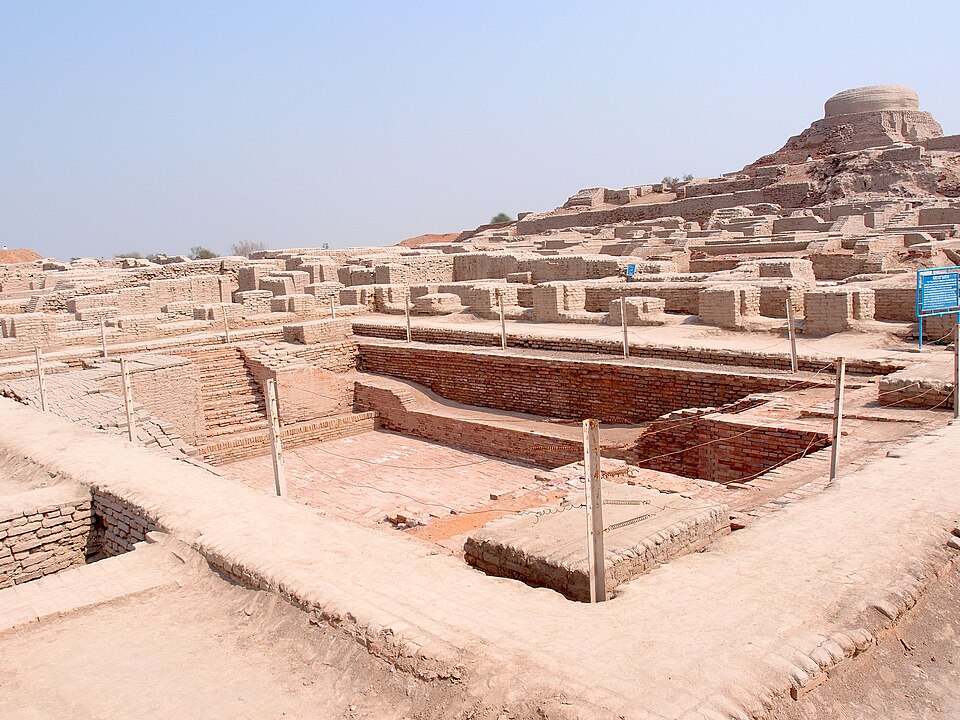 The brick-lined sunken tank of the Great Bath at Mohenjo-daro, steps descending at one end, with the excavated ruins and stupa mound of the citadel rising behind