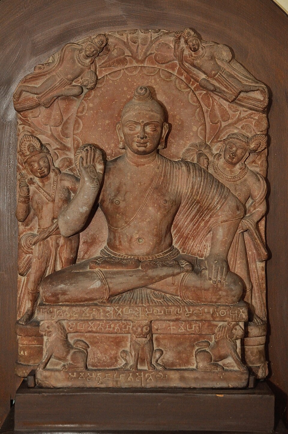Seated Buddha of the Mathura school carved in mottled red sandstone, right hand raised in abhaya mudra, flanked by attendants beneath a foliage halo, on an inscribed pedestal carved with lions