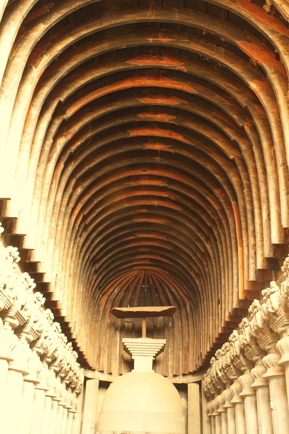 The nave of the great chaitya hall at Karla: a barrel vault ribbed with wooden beams above two colonnades of bell-capitalled pillars, closing on a rock-cut stupa crowned by a wooden umbrella