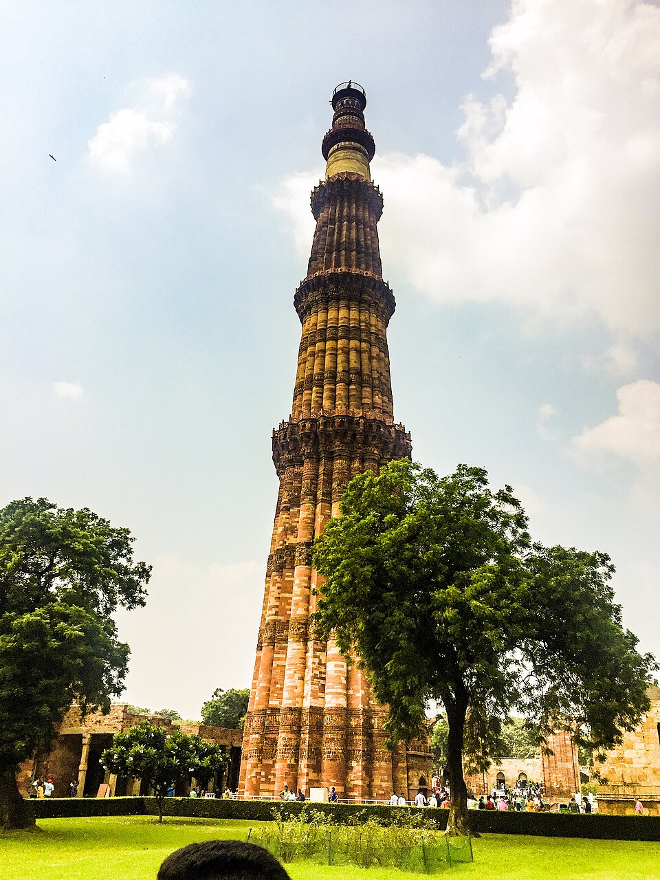 The Qutb Minar seen in full from the lawn of the complex, its five tapering storeys of fluted red sandstone separated by balconies, the upper stages faced in pale marble