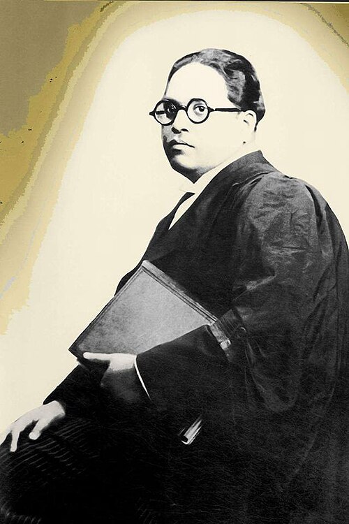 A studio portrait of B. R. Ambedkar seated in barrister's robes, wearing round spectacles and holding a large book