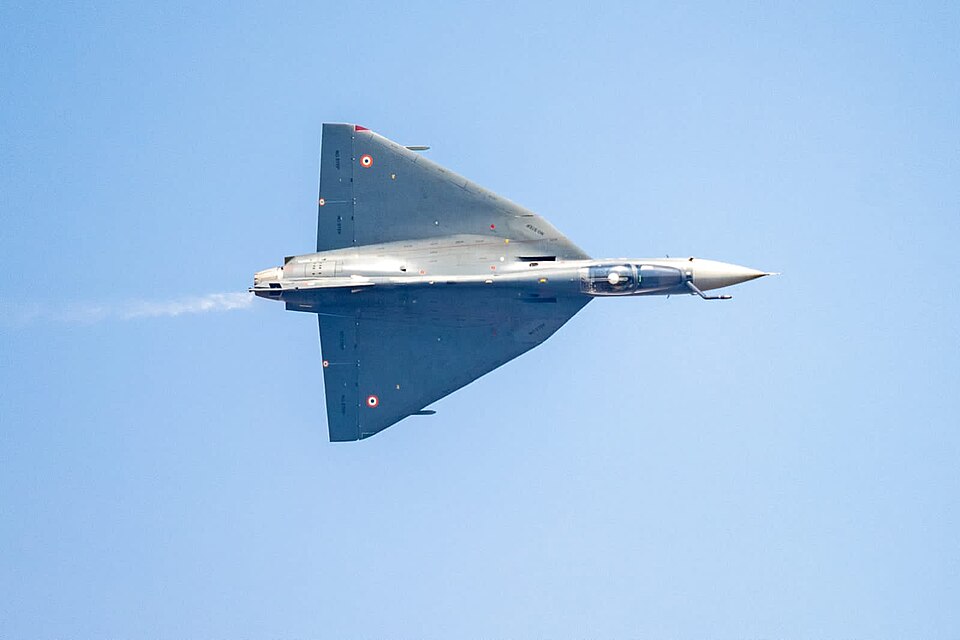 A HAL Tejas fighter banking in flight against a clear sky, its tailless delta wing plainly visible from below