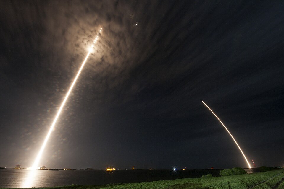 Long-exposure night photograph of a Falcon 9 launch, one bright arc rising from the pad and a second arc marking the first stage coming back to land