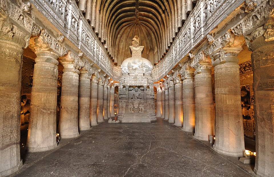 Interior of the rock-cut chaitya hall of Cave 26 at Ajanta, its ribbed vault carried on two rows of carved pillars with a stupa bearing a seated Buddha at the far end