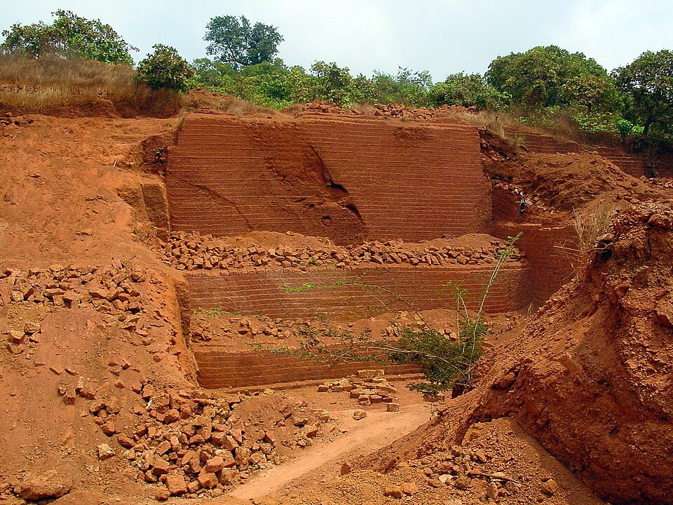 A laterite quarry face, the red rock cut back in stepped benches with saw marks and loose rectangular laterite blocks lying at the foot