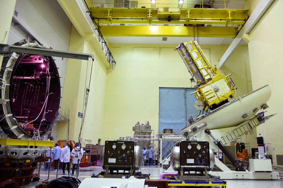 The NISAR spacecraft in a clean room at an ISRO facility, its gold-foil-wrapped body tilted on a test fixture with the long radar boom folded above it, beside the open door of a thermovacuum chamber