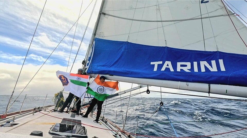 The sailing vessel Tarini under sail in a heavy Southern Ocean swell, her name lettered on the blue sail cover, with two crew on the foredeck holding the Indian tricolour and the naval ensign